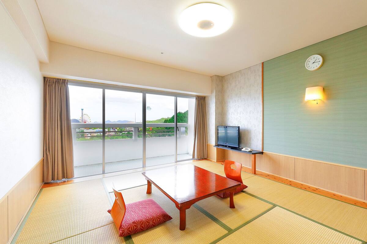 Une chambre d’hôtel avec un sol recouvert de tatamis, une porte-fenêtre coulissante donnant sur un balcon, une télévision et une horloge murale.