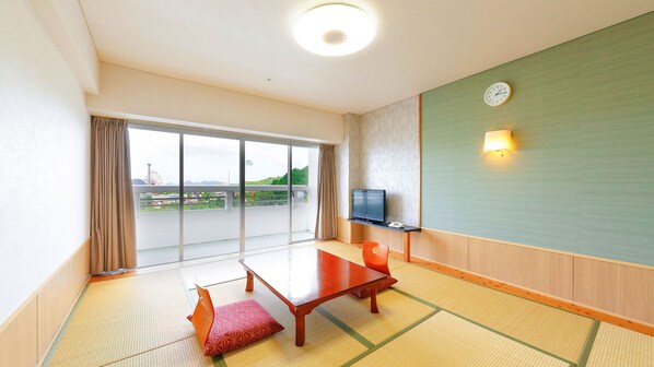 A hotel room with tatami flooring, a sliding glass door to a balcony, a television, and a clock on the wall.