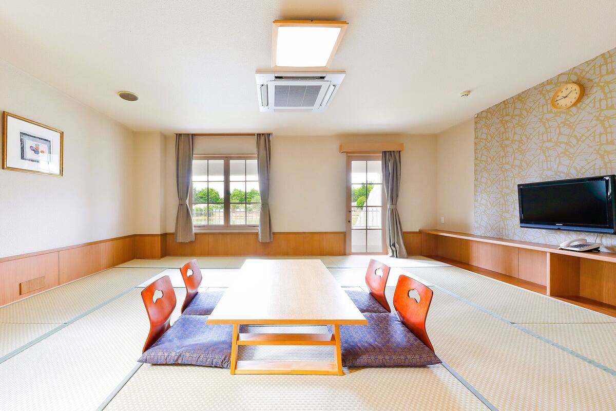 Une pièce traditionnelle avec un sol recouvert de tatamis, comprenant une longue table, des chaises, un téléviseur et une fenêtre avec des rideaux.