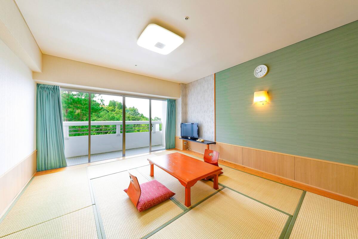 Une pièce de style japonais traditionnel, avec un sol recouvert de tatamis, une basse table en bois, un coussin rouge, un téléviseur posé sur une étagère et une porte coulissante donnant sur un balcon.