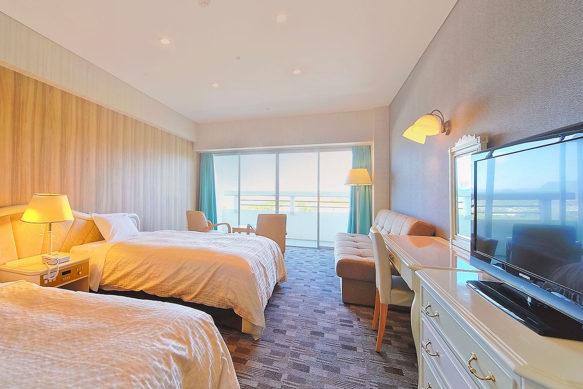 A hotel room with two beds, a TV, a desk, and a balcony with a view.