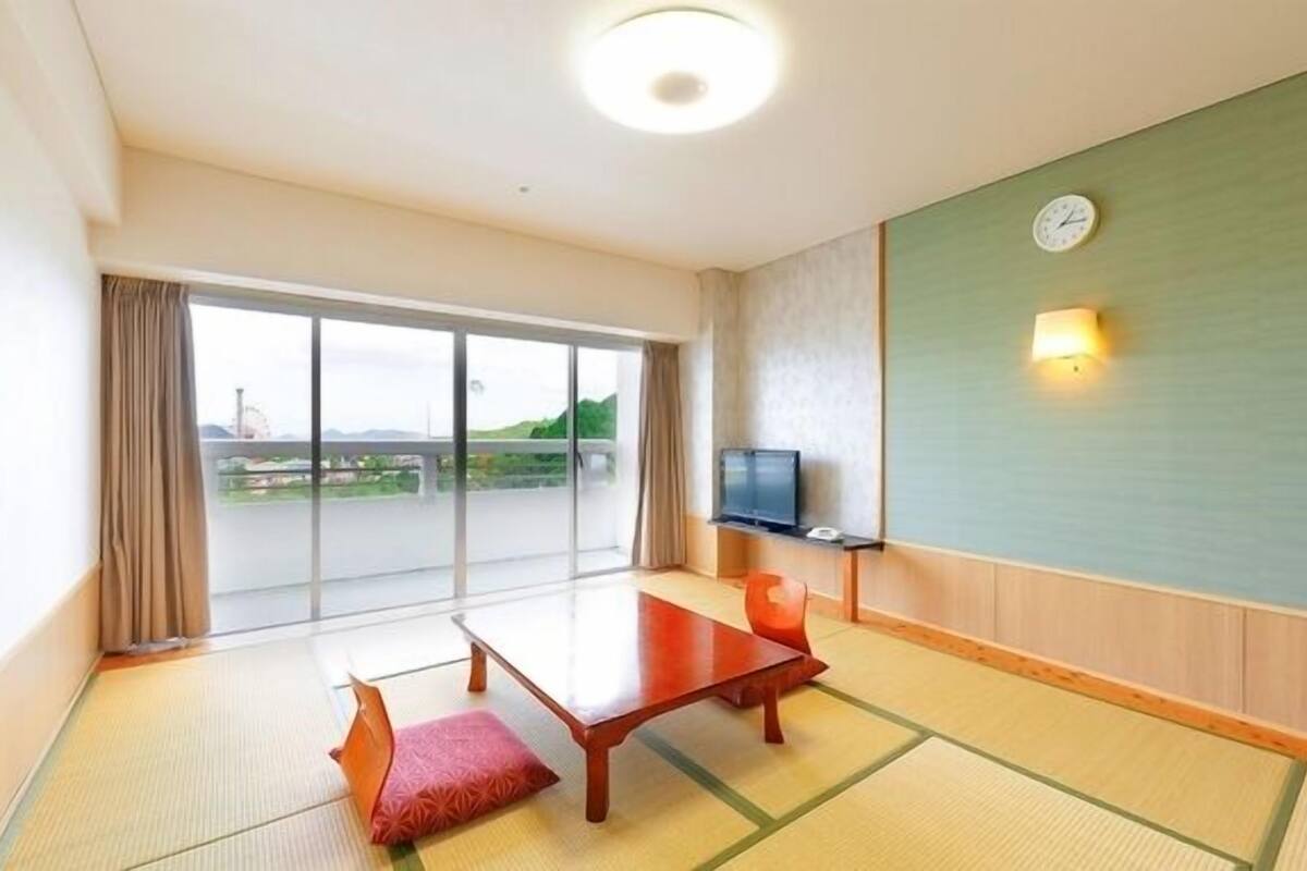 Une chambre d’hôtel avec un sol recouvert de tatamis, une porte-fenêtre coulissante donnant sur un balcon, une table en bois, un téléviseur et une horloge murale.