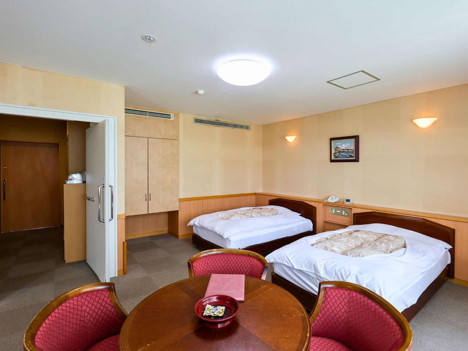 Habitación de hotel con dos camas, una mesa de comedor con sillas, una pequeña cocina americana y una puerta que da a otra habitación.