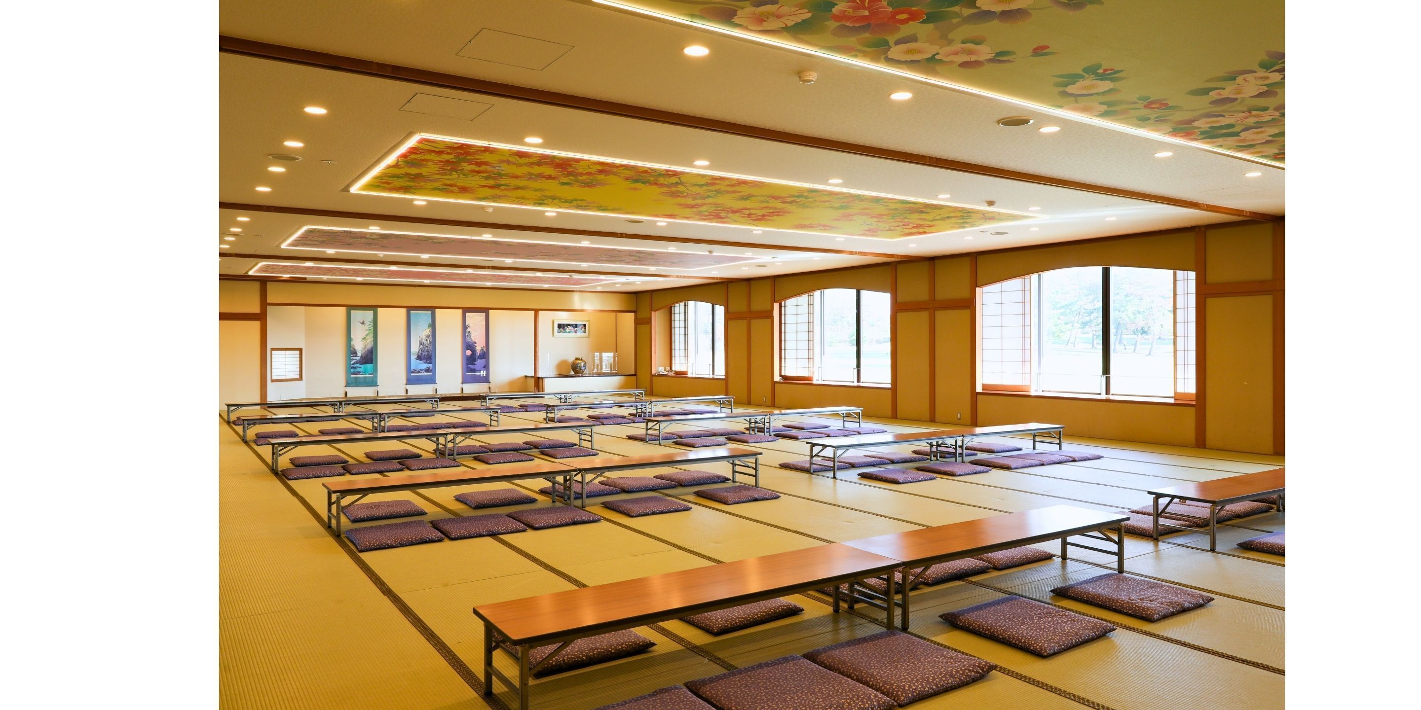 A spacious room with tatami mats, low tables, and cushions, featuring large windows and a floral ceiling design.