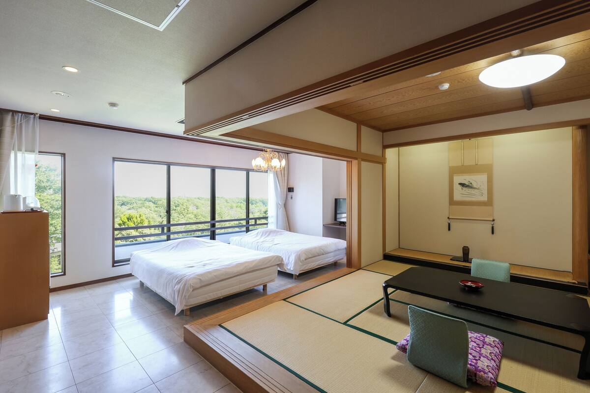 A hotel room with two beds, a dining table, tatami mats, a television, and large windows offering a view of greenery.