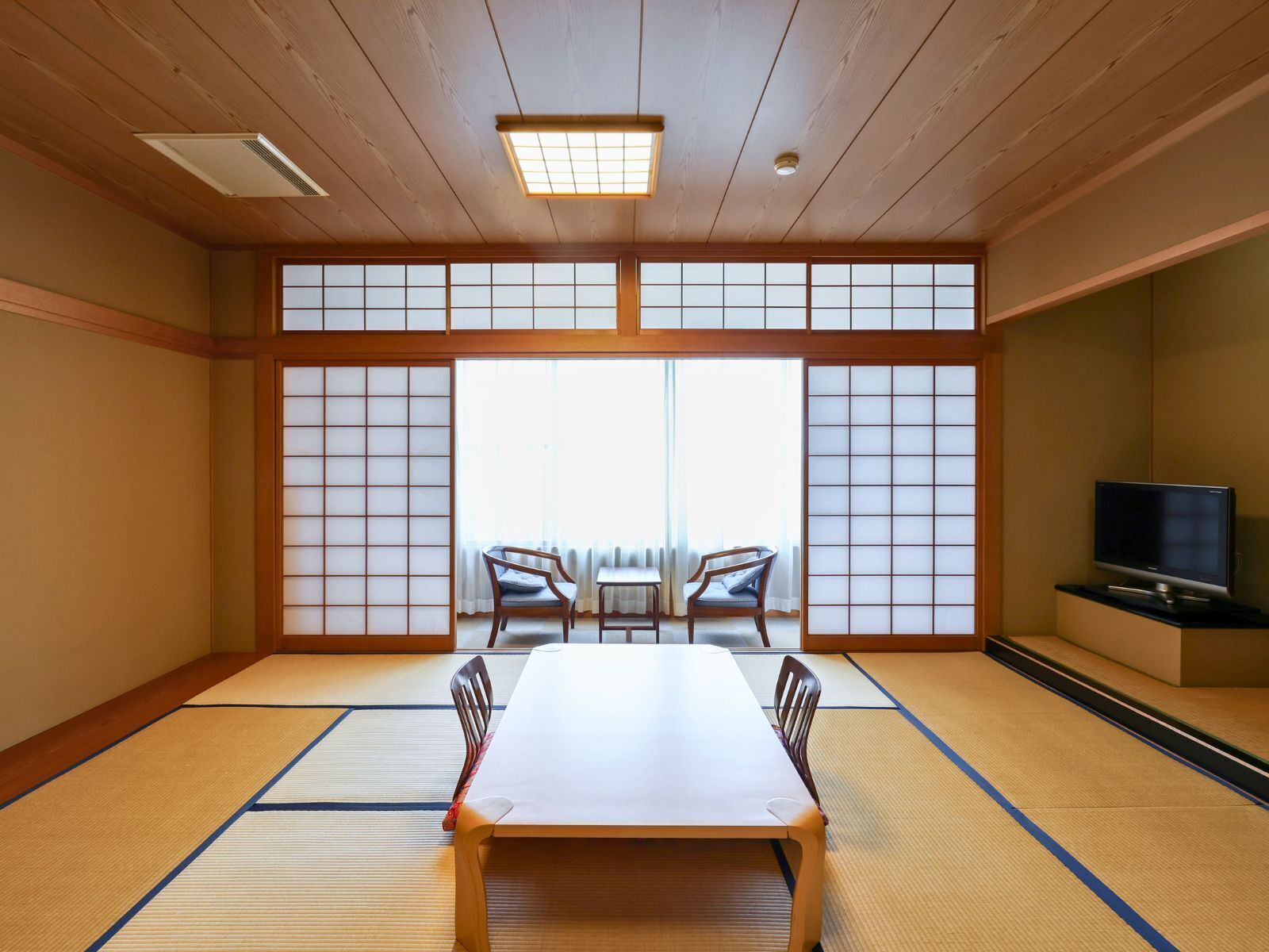 A traditional Japanese room with tatami flooring, a low table, two chairs, a television, and large shoji screens.