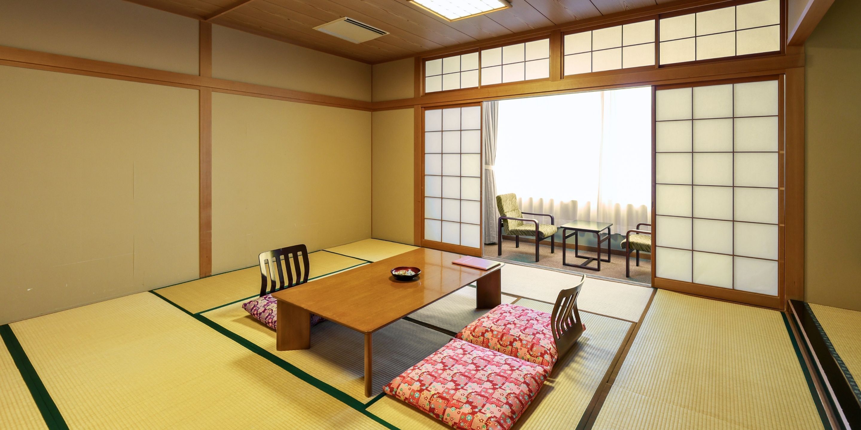 Una habitación con esteras de tatami, una mesa de madera baja, dos cojines, una mesita y una silla.
