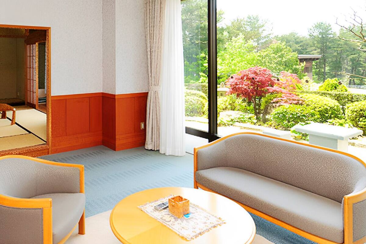 A room with a sofa, armchair, and round table, with a view of a garden through a large window.