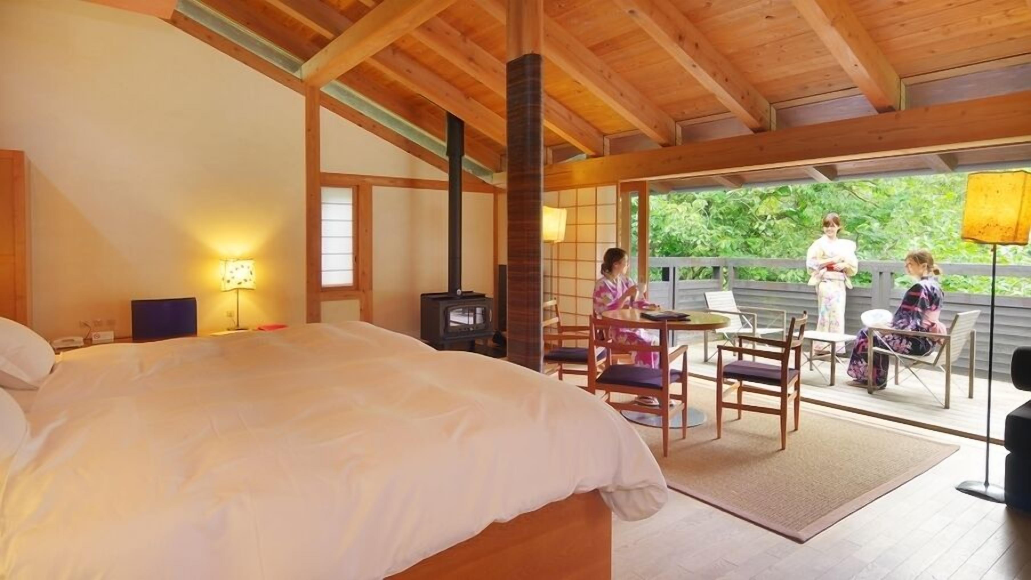 A bedroom with a bed, a wooden ceiling, a fireplace, a window, and a balcony with chairs and a table.