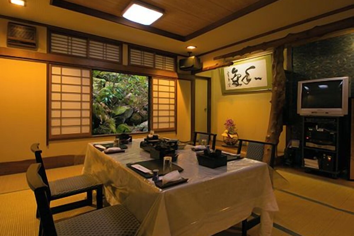 A traditional Japanese-style room with tatami flooring, a low table with cushions, a television, and a painting on the wall.
