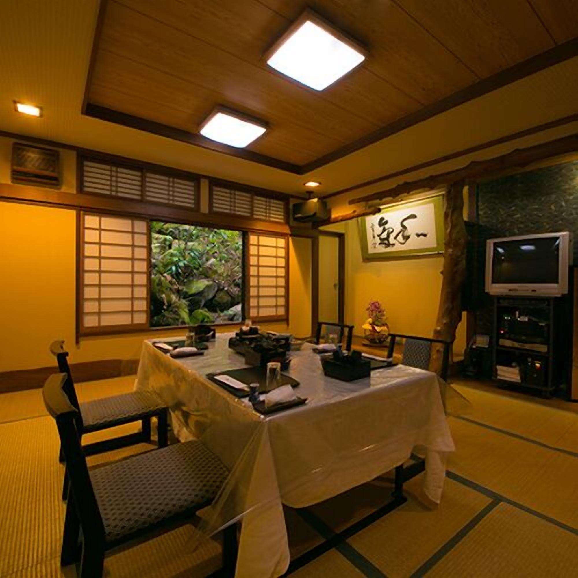Una habitación tradicional japonesa con suelo de tatami, una mesa baja con mantel blanco y un televisor sobre un soporte.