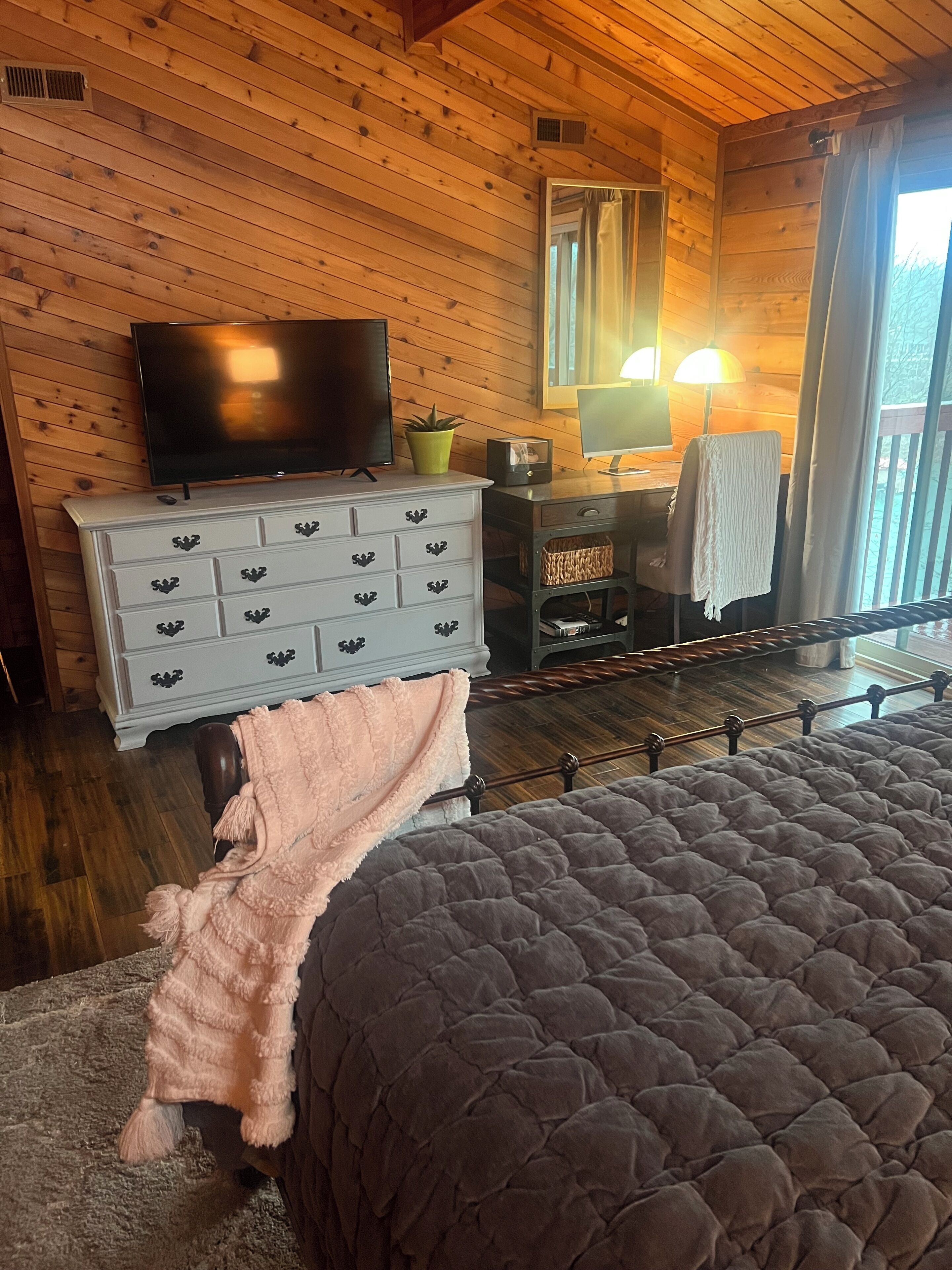 Ein Zimmer mit Holzwandverkleidung, das ein Bett, eine Kommode, einen Fernseher und einen Schreibtisch mit Lampe beinhaltet.