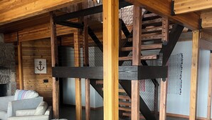 Een ruime woonkamer met een houten plafond, een stenen openhaard en een trap naar een verdieping.