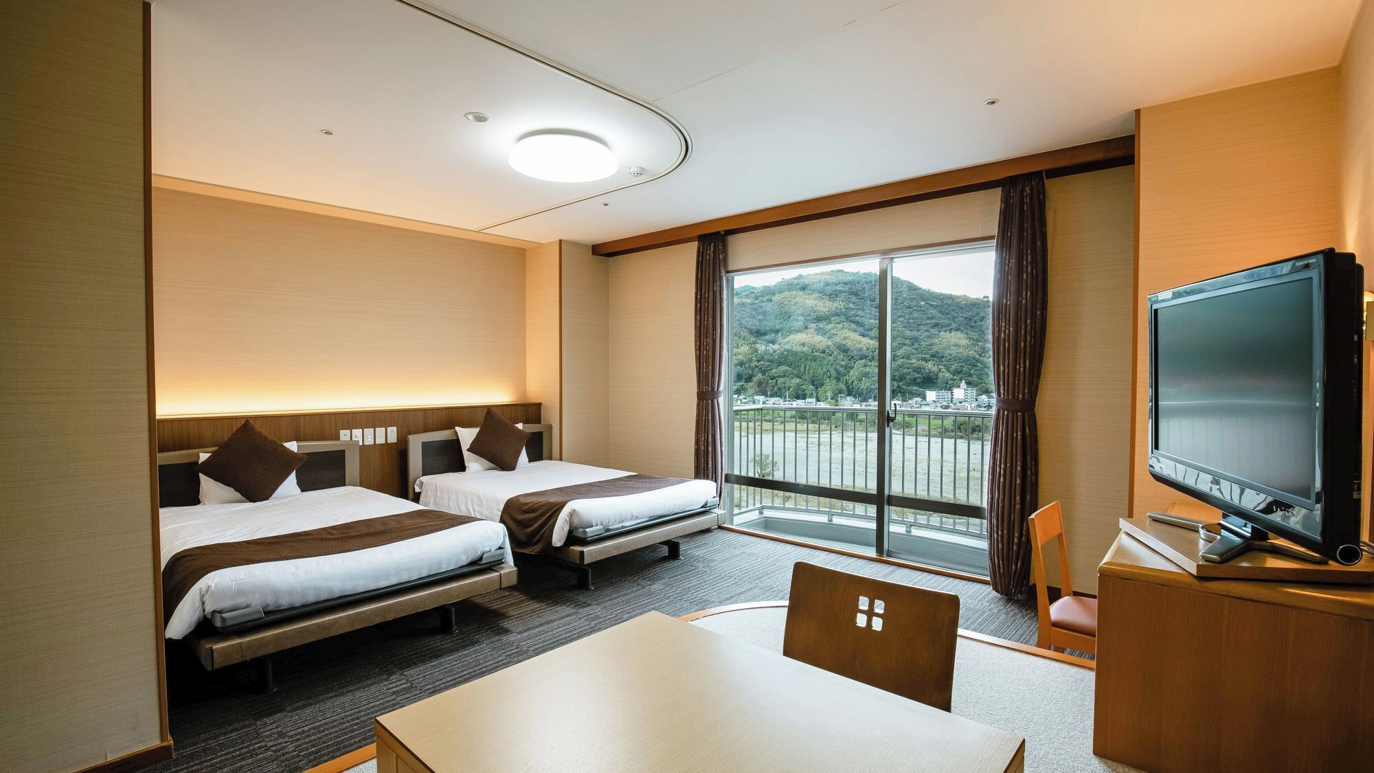 A hotel room with two beds, a TV, a table, and a view of a hilly landscape.