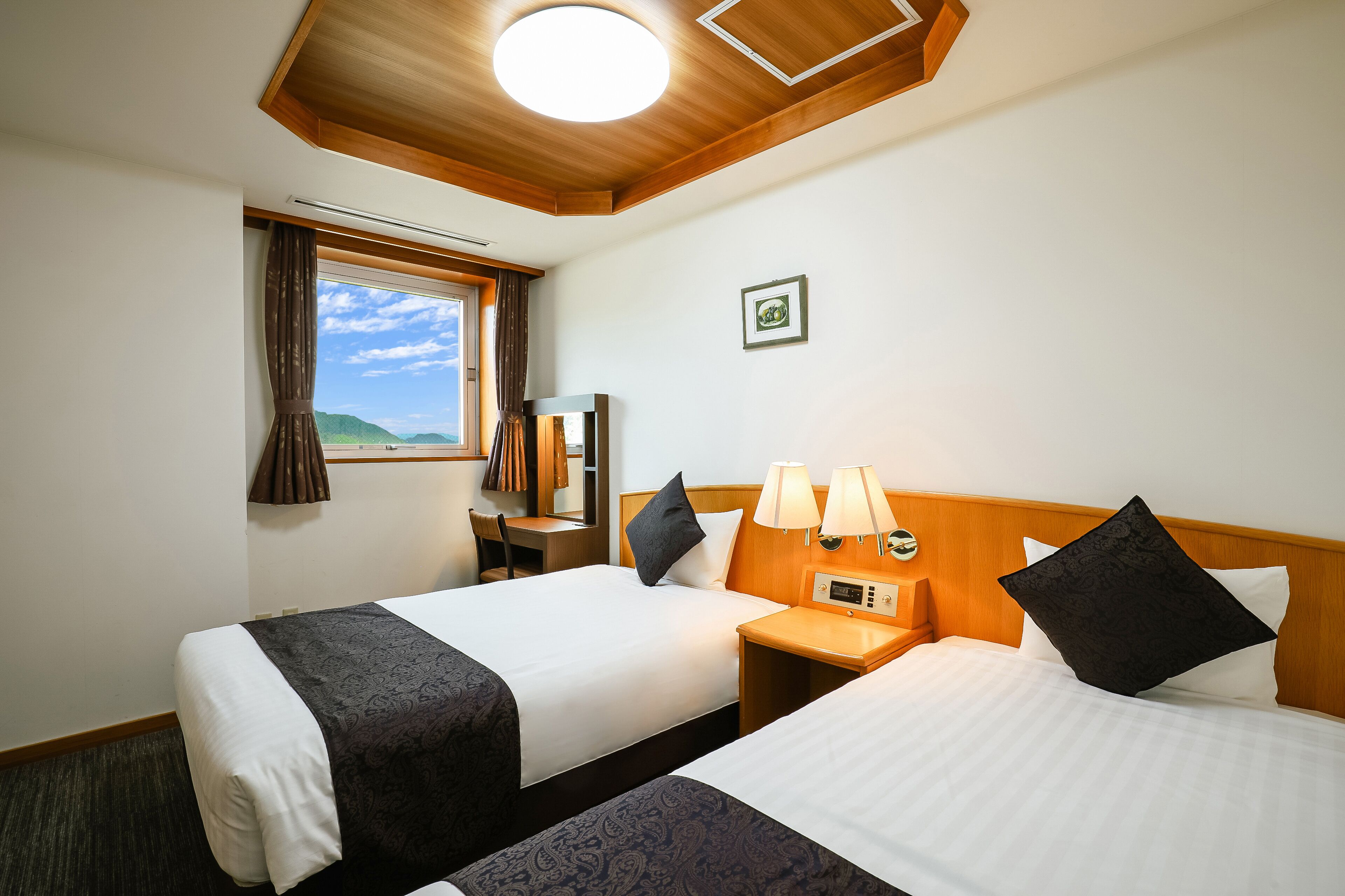 A hotel room with two beds, a desk, a chair, a window with curtains, a wall-mounted clock, and a framed picture.