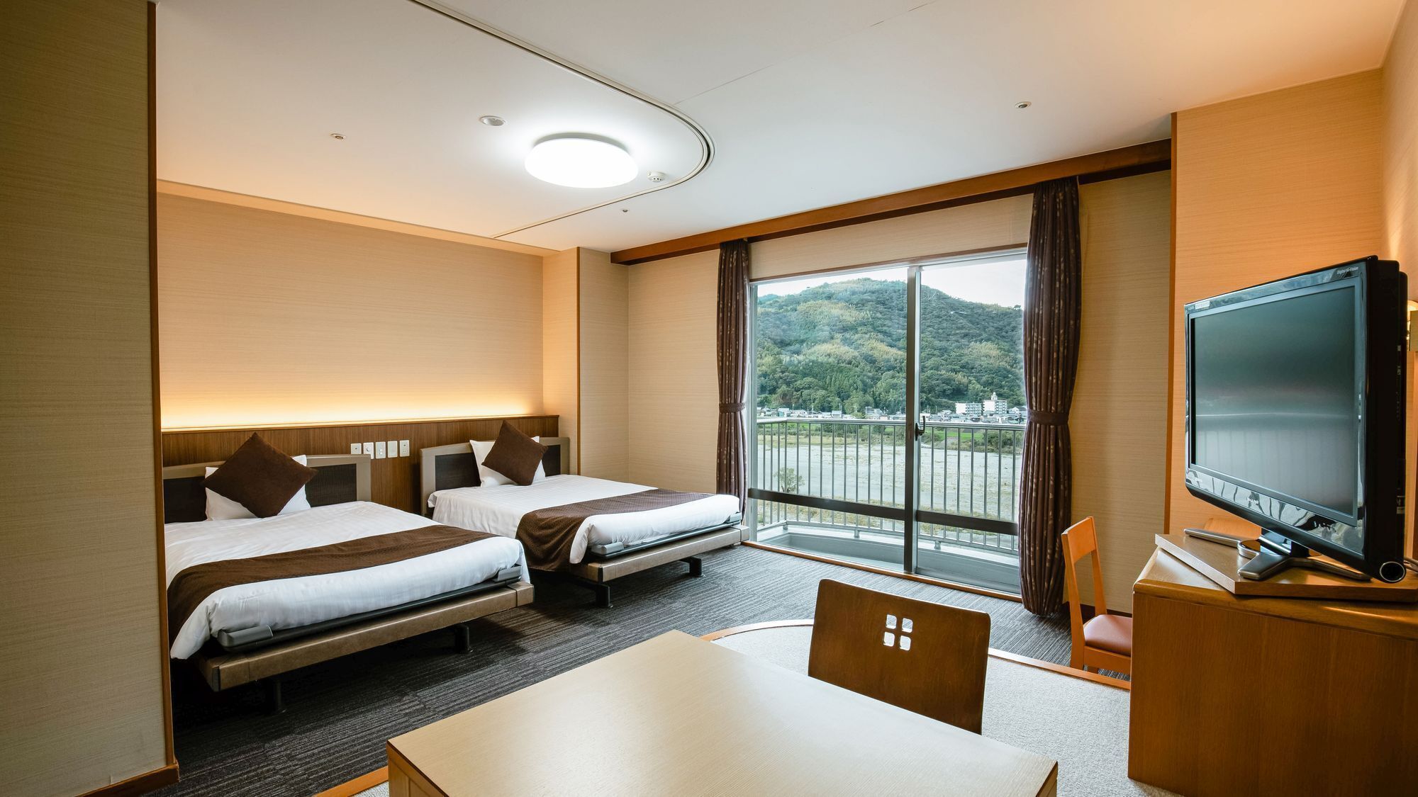 A hotel room with two beds, a TV, a table, and a view of a mountainous landscape.