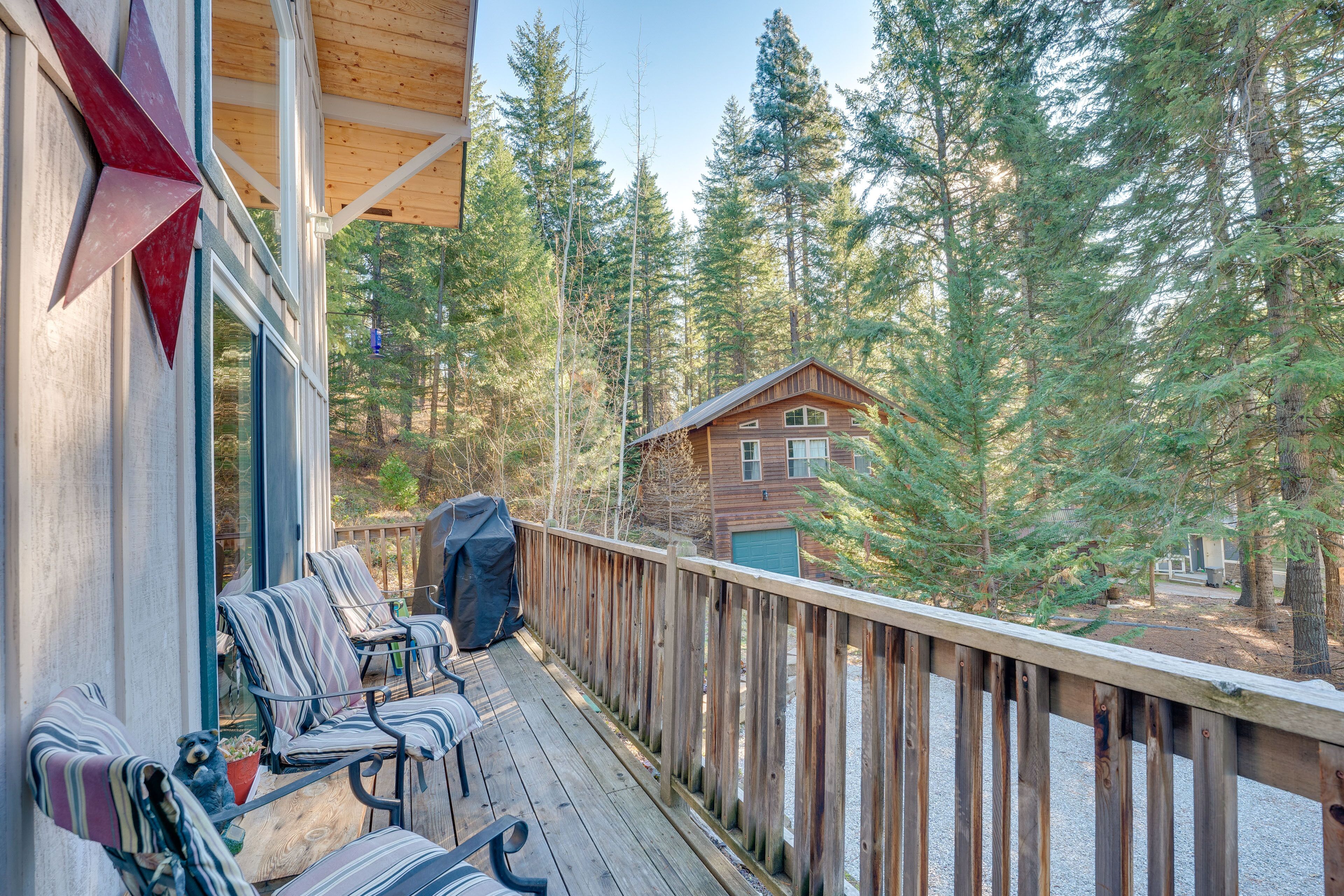 A wooden deck with chairs and a grill, overlooking a forested area with a cabin in the background.