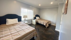 A bedroom with two beds, a window with blinds, a nightstand with a lamp, and a door.
