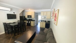 A modern living room with a dark brown sectional sofa, a wooden coffee table, and a dining area with a dark wood table and chairs. A kitchen with light-colored cabinets and a microwave is visible in the background.