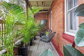 A covered outdoor area with a tiled floor, potted plants, a black chair, and a brick wall. - Renovated 3BR Heritage Home  | Private Veranda walk to the Rocks and  Barangaroo (Millers Point)