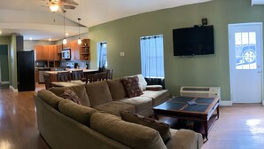 A spacious living room with a large sectional sofa, a dining area with wooden chairs, a kitchen with wooden cabinets, and a flat-screen TV mounted on the wall. - 405 S. Madison, Suite A (Watkins Glen)