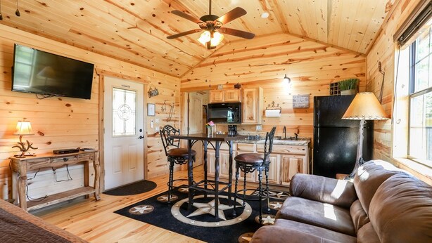 Un intérieur de chalet chaleureux avec un plafond en bois, un coin repas comprenant une table centrale et des chaises, un canapé, un téléviseur et un réfrigérateur.