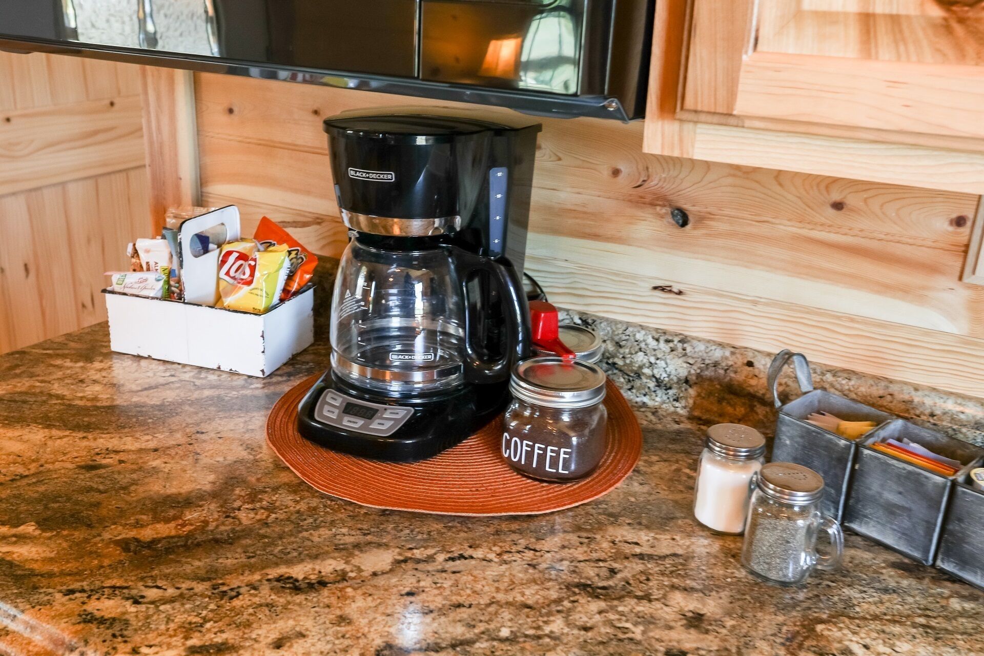 Una cocina con cafetera sobre una encimera de granito, especias en un organizador y una caja de aperitivos.