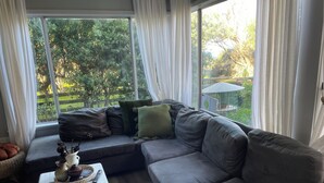 Una sala moderna con un sofá seccional gris, una mesa de centro blanca con libros y objetos decorativos, y vista a un jardín a través de amplios ventanales.