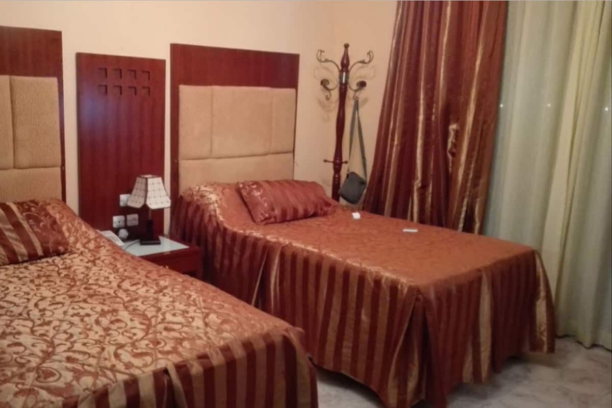 Ein Hotelzimmer mit zwei Einzelbetten, einem Nachttisch mit Lampe, einem Kleiderhaken und einem Vorhang.