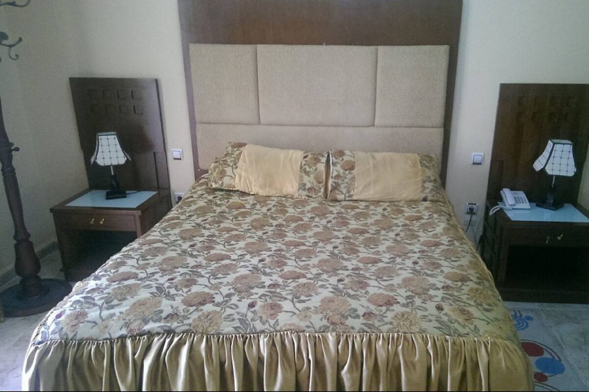Ein Hotelzimmer mit einem großen Bett, zwei Nachttischen mit Lampen, einem Holz-Kopfteil und einer blumigen Tagesdecke.