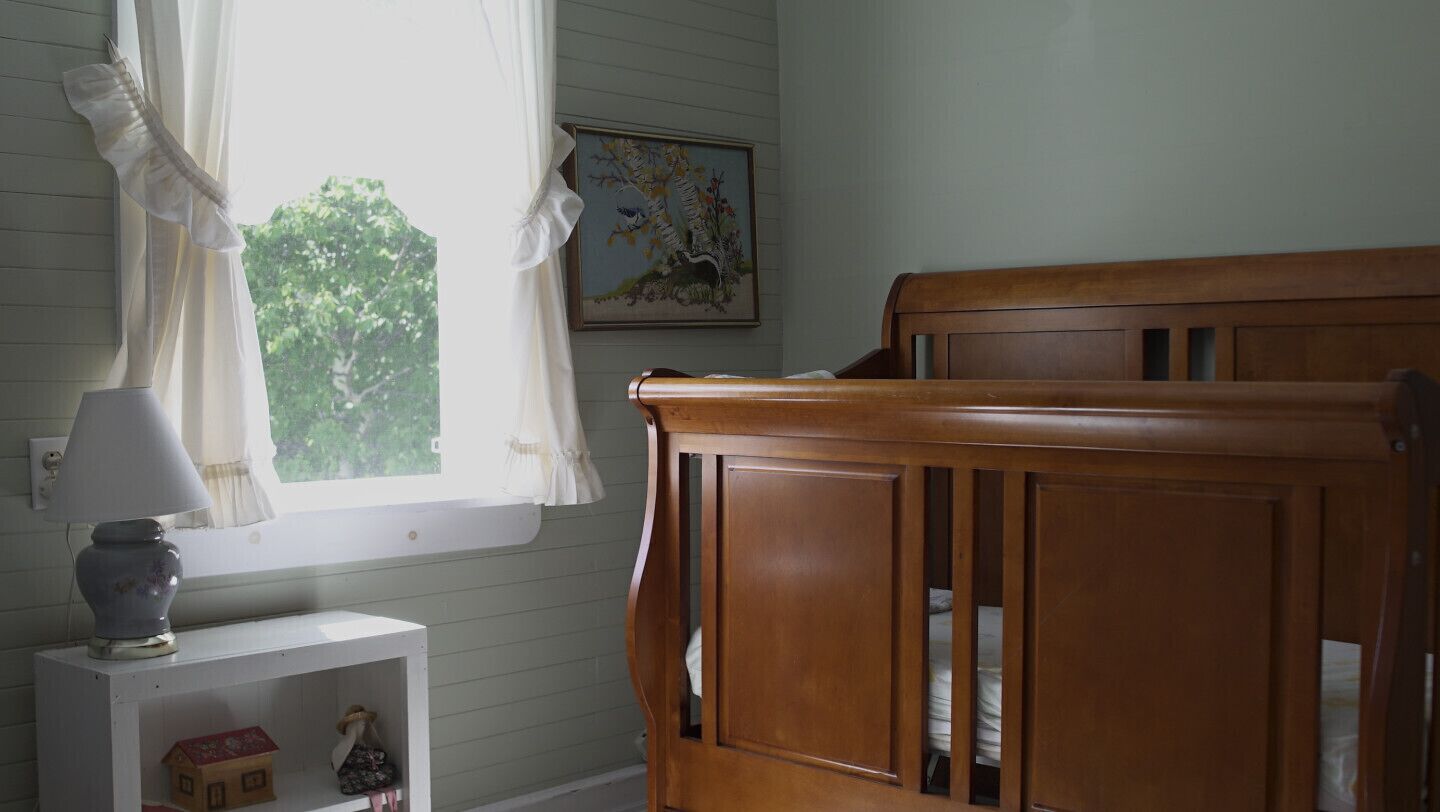 Una habitación con una cuna de madera, una mesita de noche blanca con una lámpara, un cuadro en la pared y una ventana con cortinas traslúcidas.