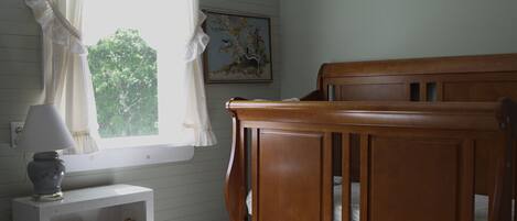 Una habitación con una cuna de madera, una mesita de noche blanca con una lámpara, un cuadro en la pared y una ventana con cortinas traslúcidas.
