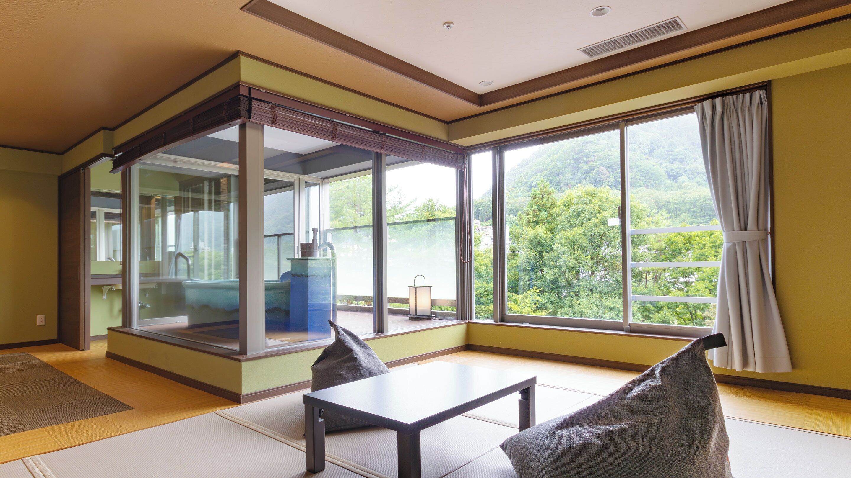 Una habitación con un gran ventanal corredera que da a un balcón, una mesa pequeña con sillas y vistas al exterior con vegetación.