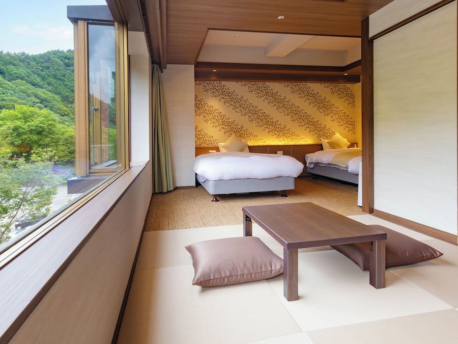 A room with a large window, a bed, a bedside table, and a view of a forested area.