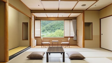 Una habitación japonesa tradicional con suelo de tatami, una mesa baja, dos sillas y vista a un bosque a través de biombos shoji.