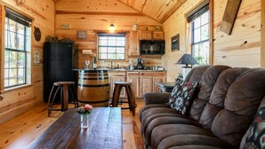 A rustic cabin interior with a wooden dining table, a sofa, a kitchen area with wooden cabinets, and a large window. - God's Country Cabin- Faith I Country Retreat (Fredericksburg)