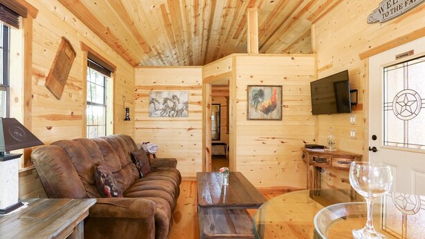 A cozy wooden cabin interior with a sofa, a coffee table, a TV, and a glass table with wine glasses. - God's Country Cabin- Faith I Country Retreat (Fredericksburg)