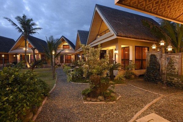 Un resort con varias construcciones tipo bungalow, cada una con techo de paja y rodeada de jardines ajardinados.