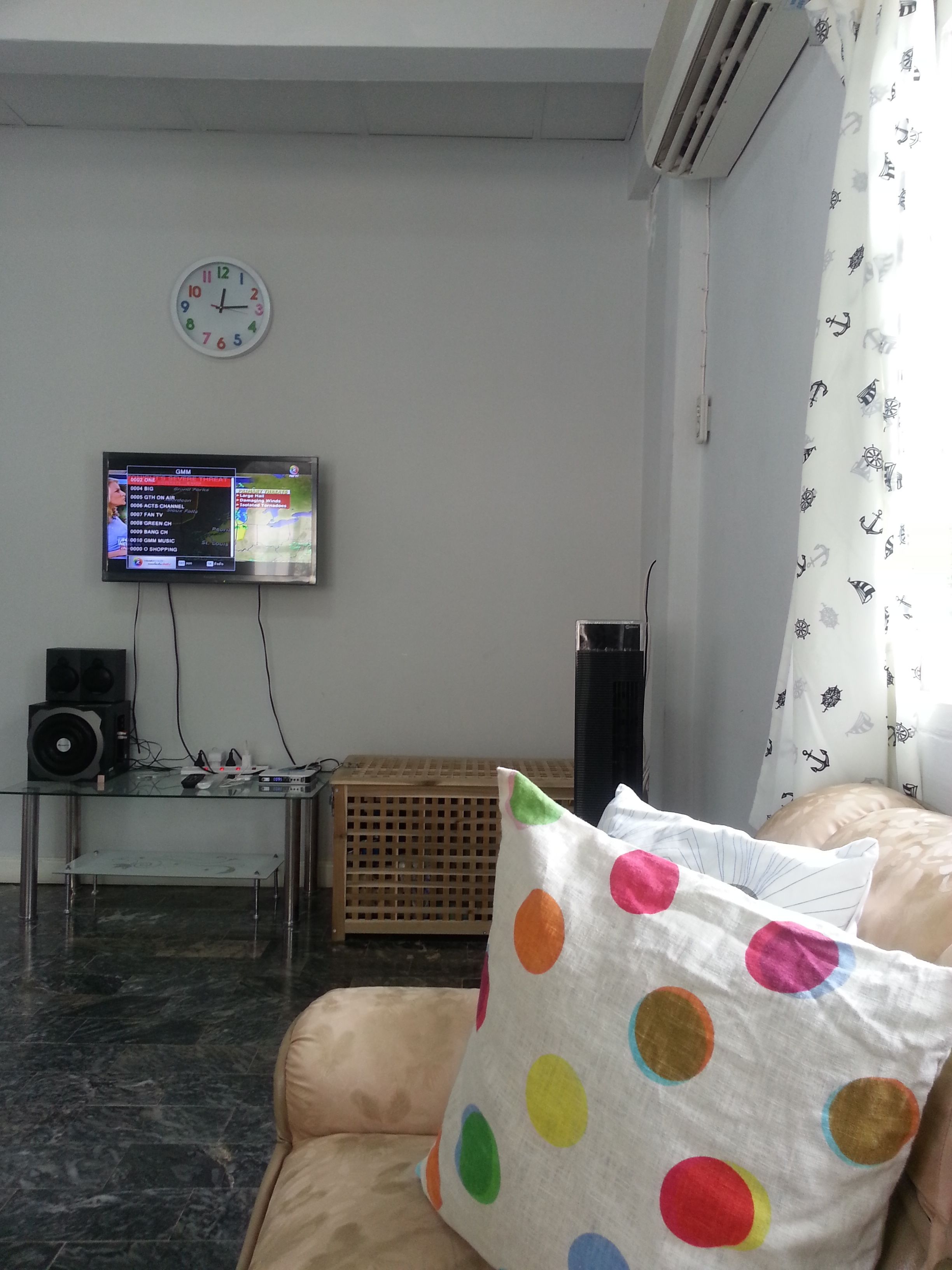 Una sala de estar con un sofá, un televisor montado en la pared, un reloj y un altavoz sobre una mesa.