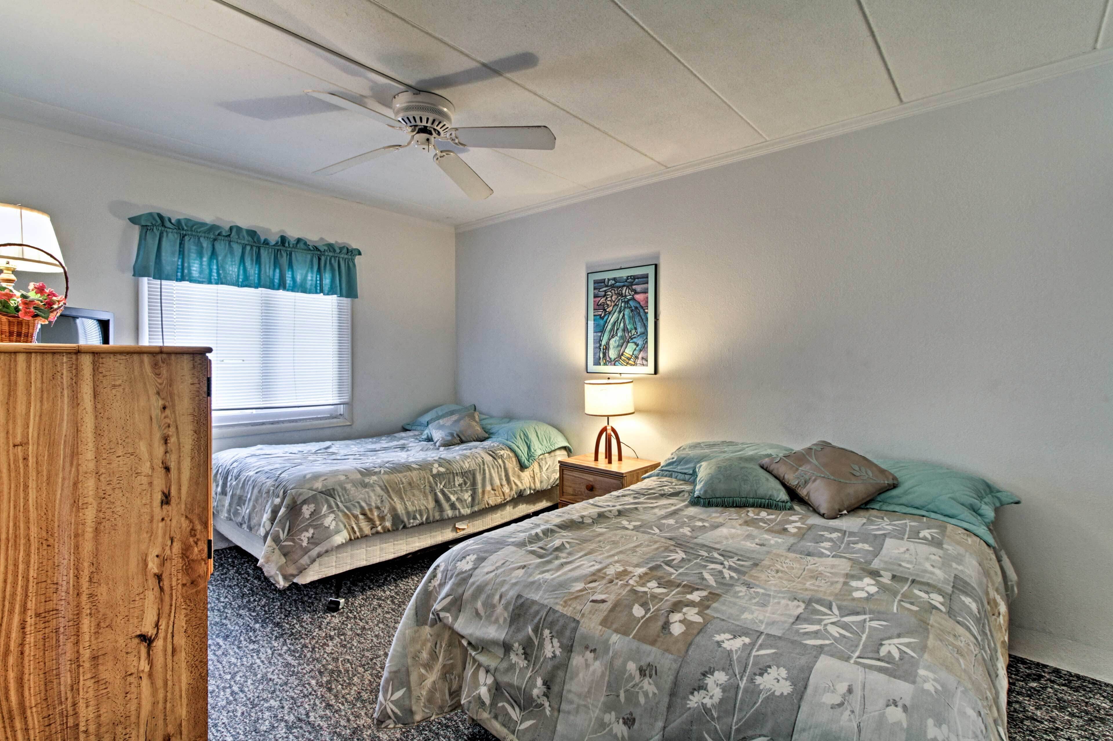 Une chambre avec deux lits, un ventilateur de plafond, une fenêtre avec des stores, une lampe et un tableau encadré au mur.