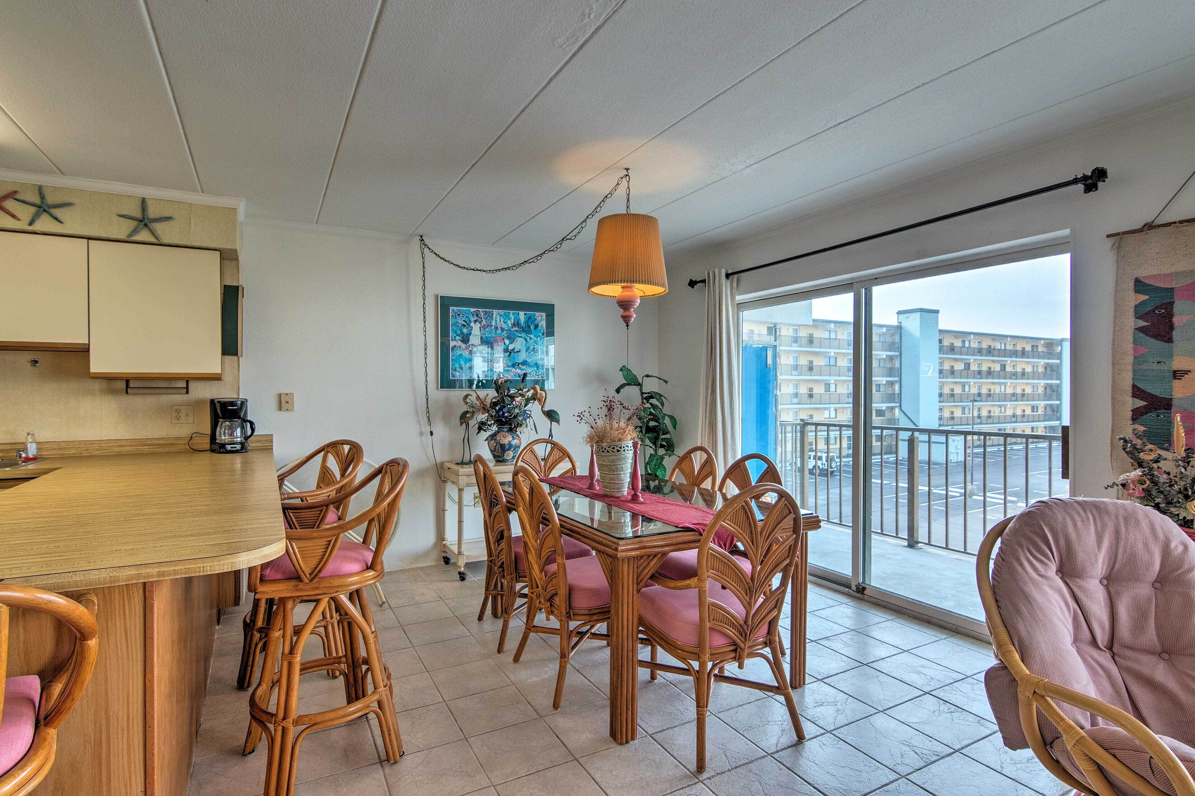 A dining area with a round table and six wicker chairs, a hanging lamp, and a view of buildings through large windows.