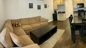 A living room with a sofa, a coffee table, and a dining area with a table and chairs. There is a kitchen in the background with cabinets and appliances. - Watkins Glen 3-Bedroom Suites Unit 4 (Watkins Glen)