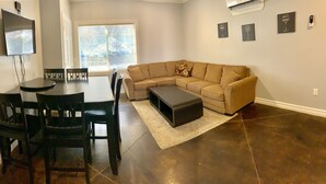 A living room with a sectional sofa, a dining table with chairs, a flat-screen TV mounted on the wall, and a window with blinds. - Watkins Glen 3-Bedroom Suites Unit 4 (Watkins Glen)