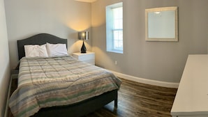 A bedroom with a bed, nightstand, lamp, window, and a desk. - 405 S. Madison, Suite B (Watkins Glen)