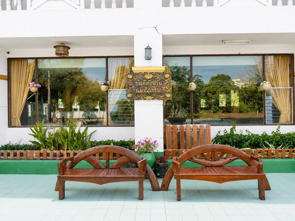 A wooden bench with a sign in front, a white building with large windows, and a view of greenery. - Royal Jommanee Hotel (Nong Khai)