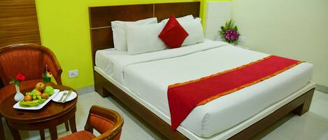 A hotel room with a bed, a dining table with fruit and a vase, and a chair.