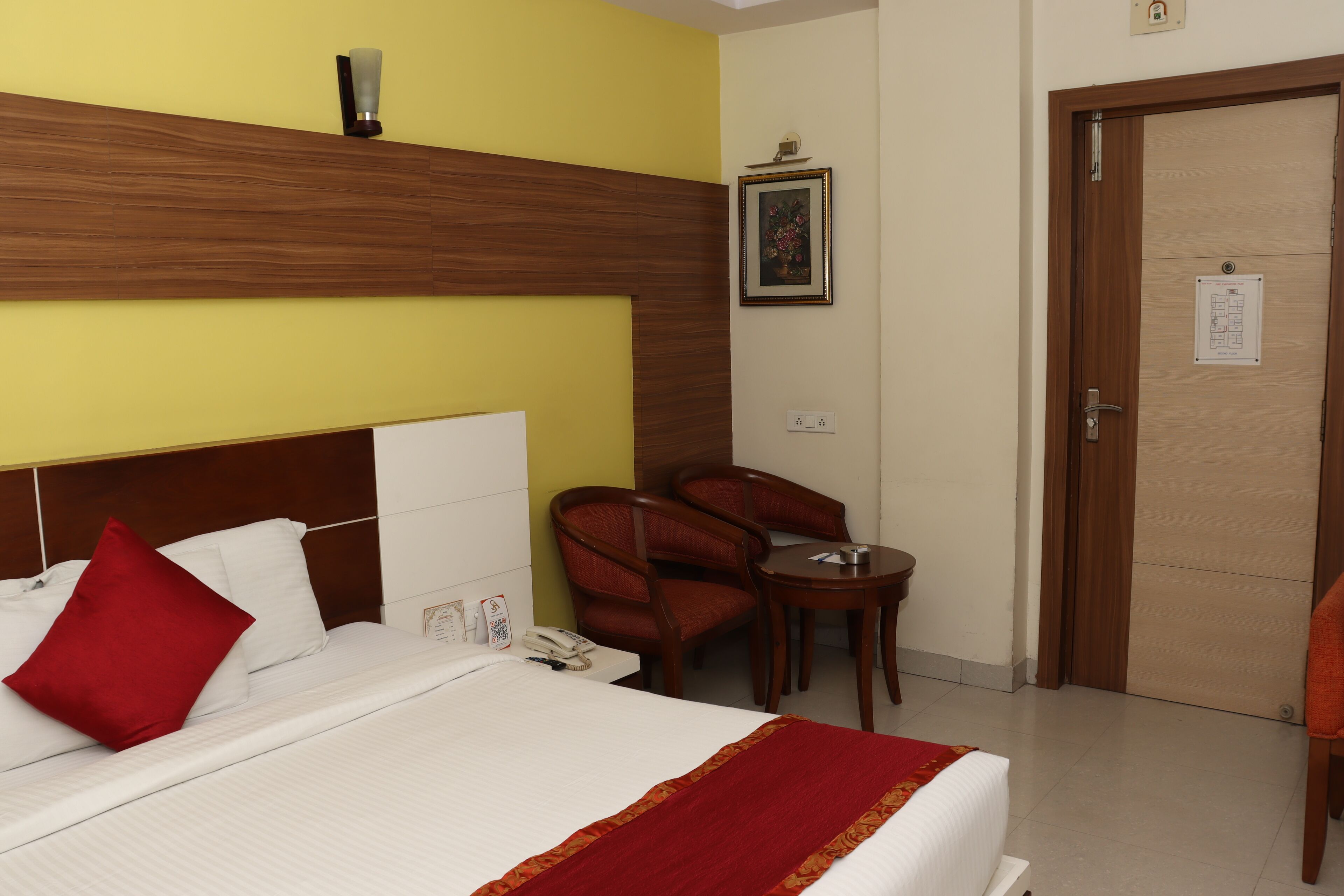 A hotel room with a bed, a wooden headboard, a red pillow, a small table, and a chair.