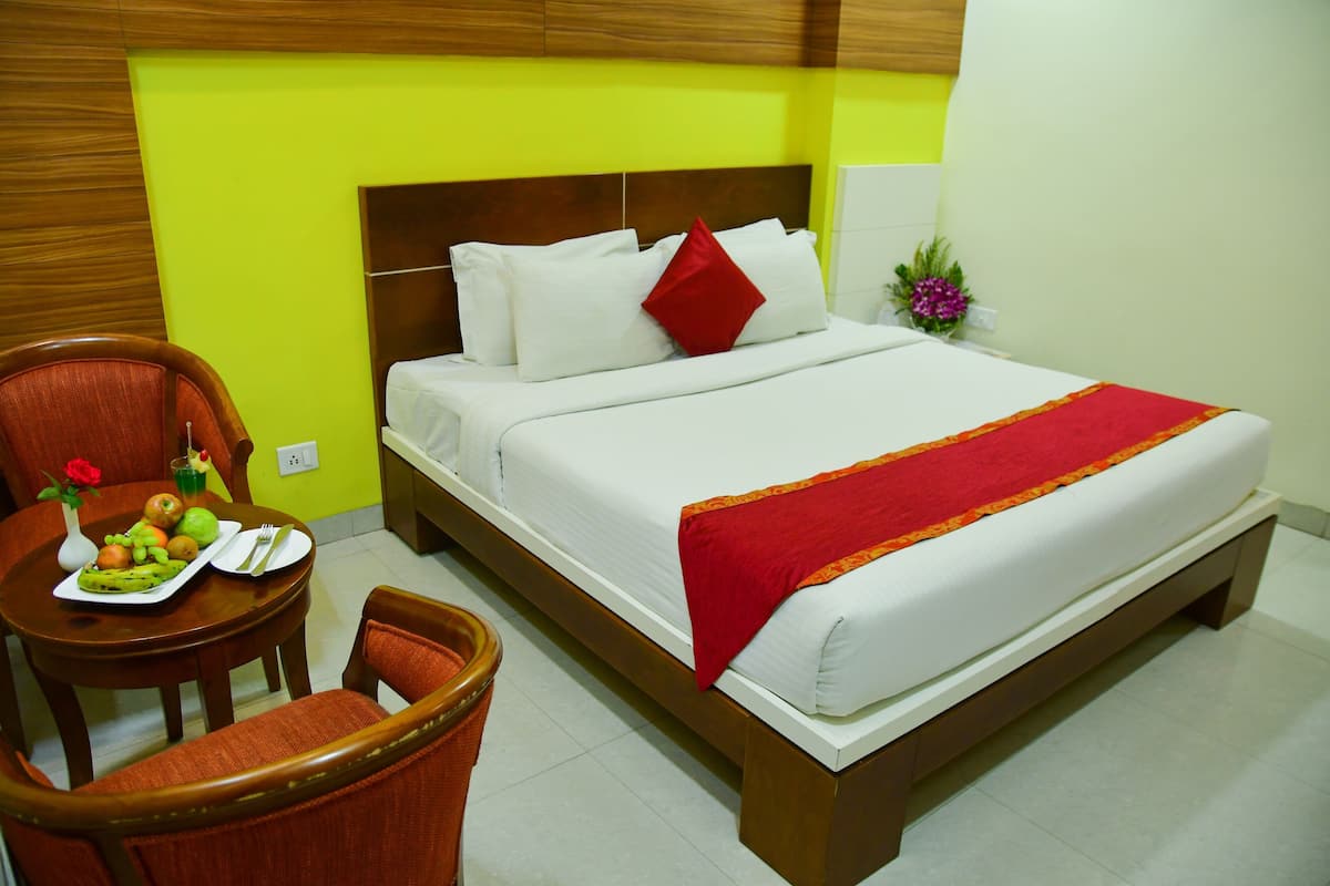 Quarto de hotel com uma cama, uma mesa de jantar com frutas, uma cadeira e uma pequena mesa com um vaso de flores.
