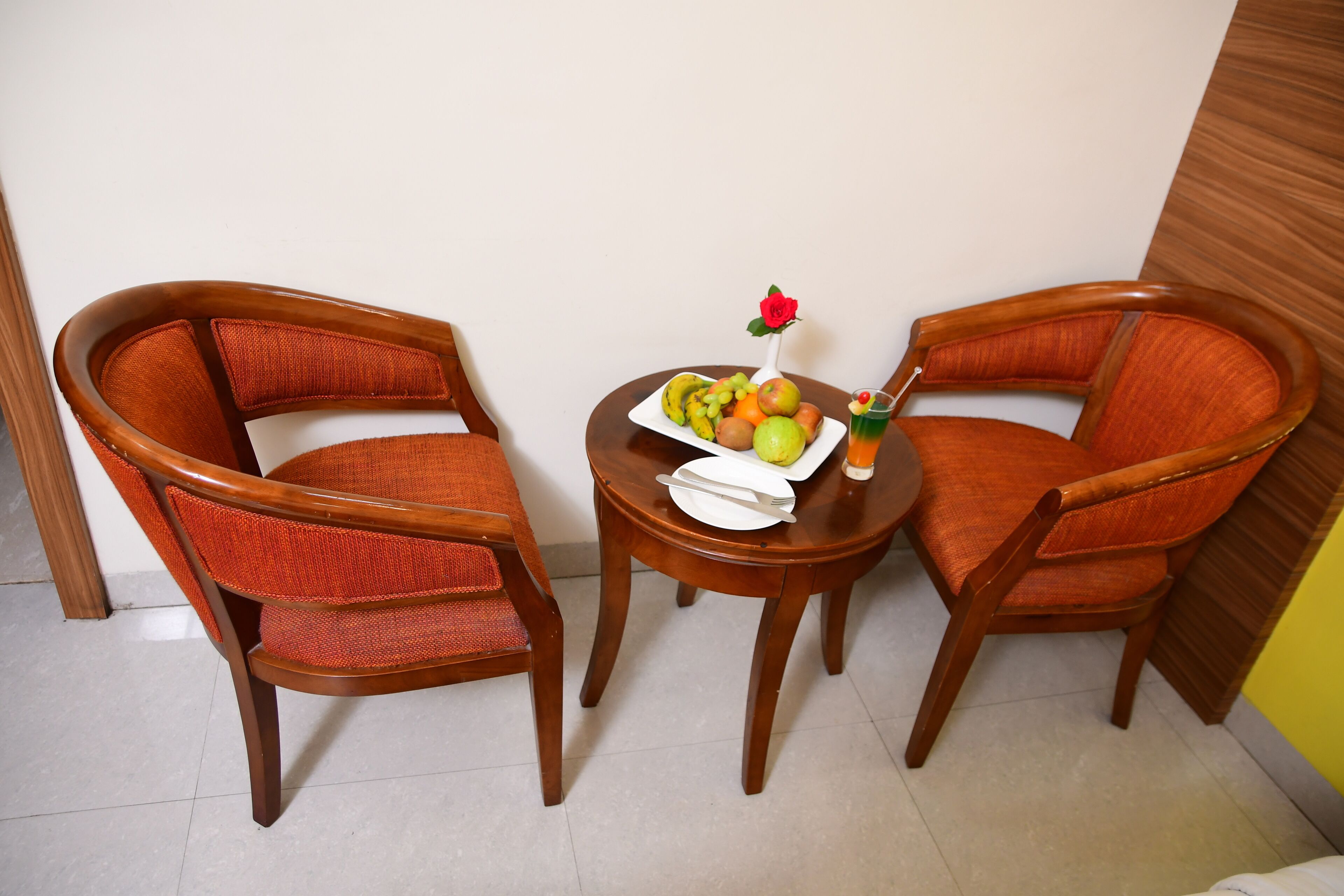 Two wooden chairs with red cushions, a small round table with a fruit plate, a white plate, and a rose in a vase.