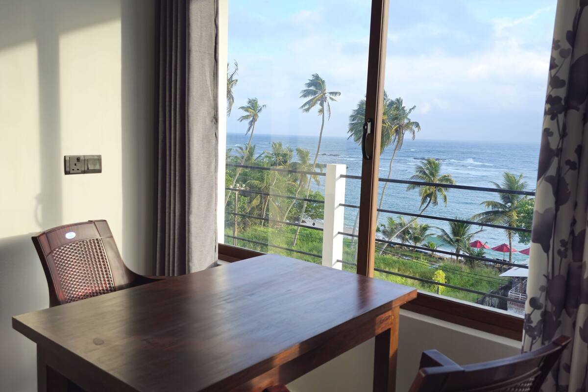 A balcony with a wooden table and chairs, overlooking a beach with palm trees and the ocean.